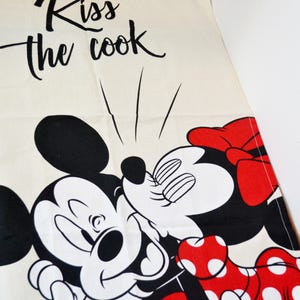 May include: A cream-colored kitchen towel featuring a black graphic of Mickey Mouse kissing Minnie Mouse. Minnie wears a red bow and a red dress with white polka dots. The text "Kiss the cook" is printed in black script above the characters.