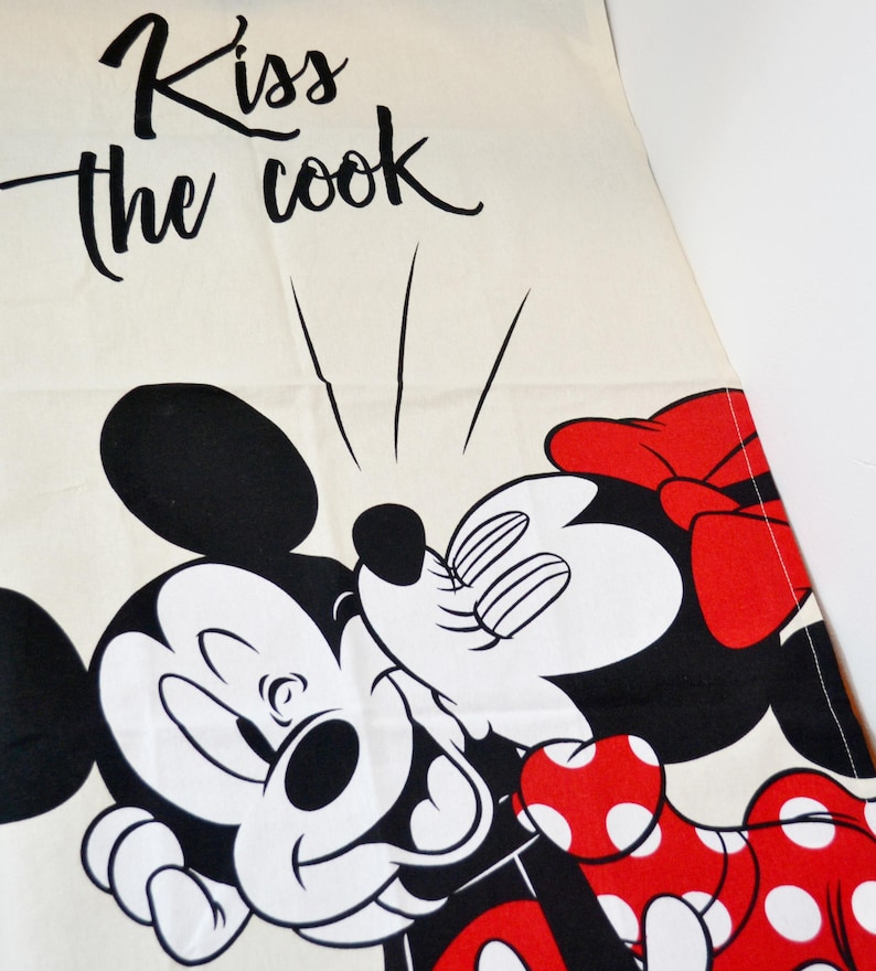 May include: A cream-colored kitchen towel featuring a black graphic of Mickey Mouse kissing Minnie Mouse. Minnie wears a red bow and a red dress with white polka dots. The text "Kiss the cook" is printed in black script above the characters.