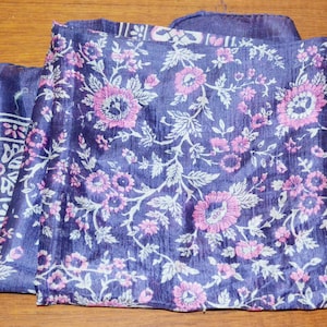 May include: A folded square scarf in a deep purple hue featuring a repeating floral pattern in shades of light pink and white. The fabric displays a subtle sheen and a visible weave, with a distinct border design along the left edge of the textile.