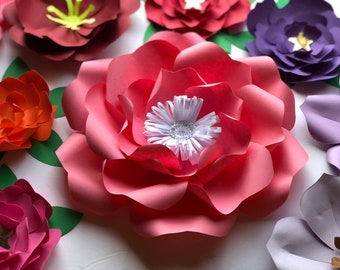 May include: A large, pink paper flower with a white center. The flower is made of multiple layers of paper and has a ruffled edge. The flower is surrounded by other paper flowers in various colors, including orange, purple, and red.