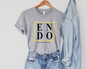 Puede incluir: Camiseta gris con un gráfico negro que dice "ENDO" en un cuadro amarillo. El texto "ENDOMETRIOSIS" está escrito debajo del cuadro. La camiseta está en una percha y se combina con una chaqueta vaquera azul claro y pantalones cortos de mezclilla.