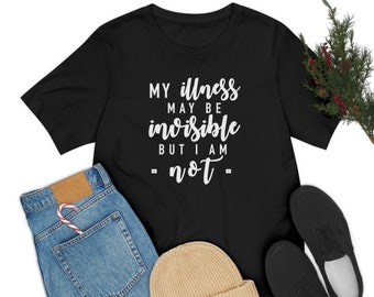 May include: Black t-shirt with white text that reads "My illness may be invisible but I am not". The shirt is laid flat on a white background with a pair of blue jeans, a brown beanie, and black sneakers.