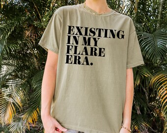 Op de afbeelding: Een groen T-shirt met de tekst "EXISTING IN MY FLARE ERA." in het zwart gedrukt.