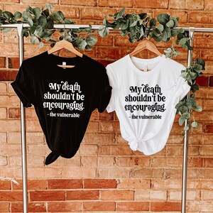 May include: Two t-shirts with the same text printed on them. The text reads "My death shouldn't be encouraging. - the vulnerable". One t-shirt is black and the other is white.