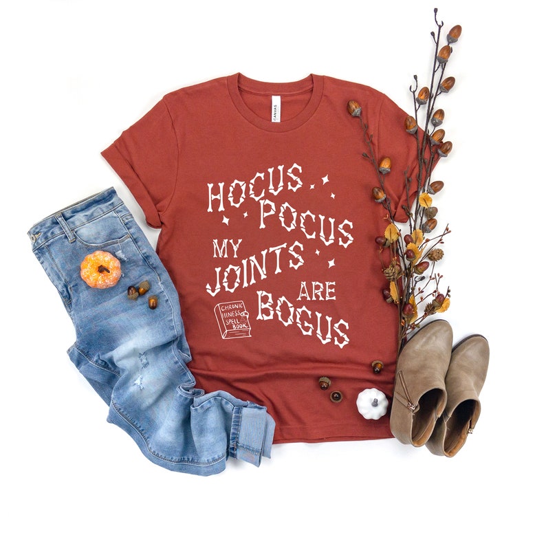May include: A rust-colored t-shirt with the text "Hocus Pocus My Joints Are Bogus" in white. The shirt is styled with blue jeans, brown boots, and fall-themed decor.