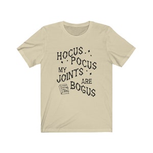 May include: A beige t-shirt with black text that reads "Hocus Pocus My Joints Are Bogus" and a small illustration of a book with the text "Chronic Pain Spell Book".
