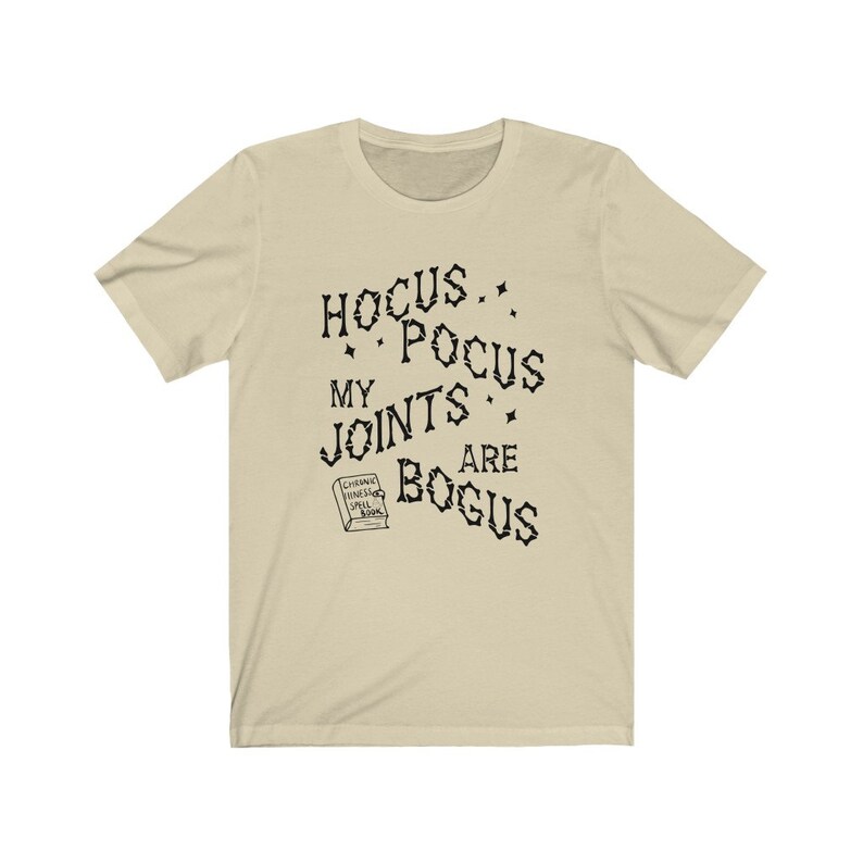 May include: A beige t-shirt with black text that reads "Hocus Pocus My Joints Are Bogus" and a small illustration of a book with the text "Chronic Pain Spell Book".