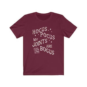 May include: Burgundy t-shirt with white text that reads "Hocus Pocus My Joints Are Bogus" and a small image of a book that says "Chronic Haxing Spell Book".