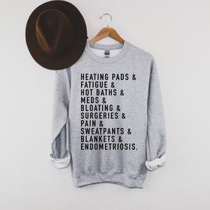 May include: Gray sweatshirt with black text that reads "HEATING PADS & FATIGUE & HOT BATHS & MEDS & BLOATING & SURGERIES & PAIN & SWEATPANTS & BLANKETS & ENDOMETRIOSIS."