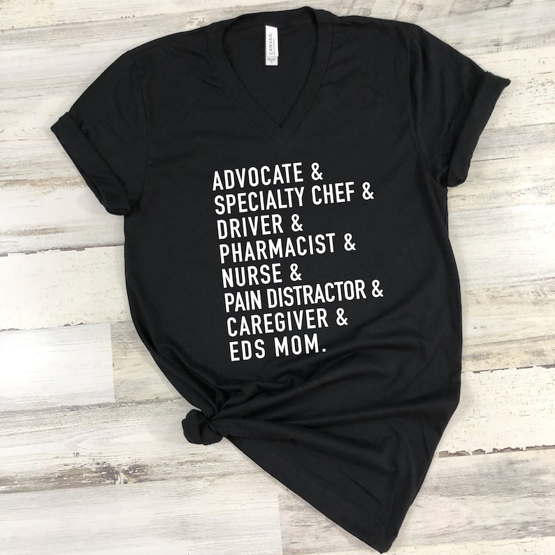 以下が含まれることがあります： 白い文字で「ADVOCATE & SPECIALTY CHEF & DRIVER & PHARMACIST & NURSE & PAIN DISTRACTOR & CAREGIVER & EDS MOM.」と書かれた、黒いVネックTシャツ