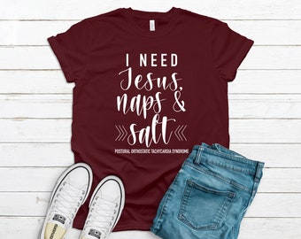 May include: A maroon t-shirt with white text that reads "I need Jesus, naps & salt" with the acronym "POTS" (Postural Orthostatic Tachycardia Syndrome) below. The shirt is laid out on a white wooden surface with a pair of white sneakers and blue jeans.