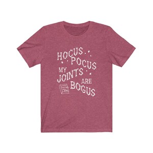May include: A heather red t-shirt with a white graphic that says "Hocus Pocus My Joints Are Bogus" with a small book graphic that says "Chronic Pain Spell Book".
