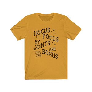 May include: A mustard yellow t-shirt with black text that reads "Hocus Pocus My Joints Are Bogus". A small black and white illustration of a book with the text "Chilling Vines Spell Book" is below the text.