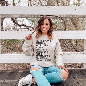 May include: A light beige sweatshirt with black text that reads "Heating pads & fatigue & hot baths & meds & bloating & surgeries & pain & sweatpants & blankets & endometriosis."