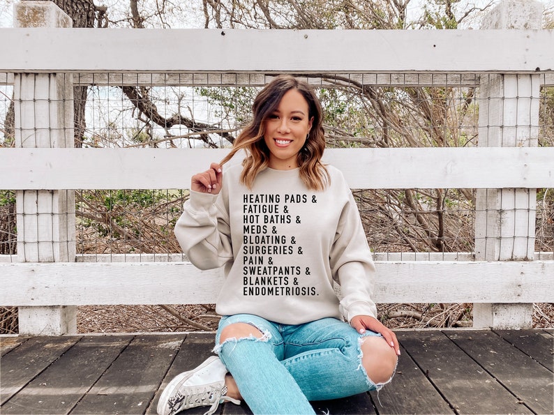May include: A light beige sweatshirt with black text that reads "Heating pads & fatigue & hot baths & meds & bloating & surgeries & pain & sweatpants & blankets & endometriosis."