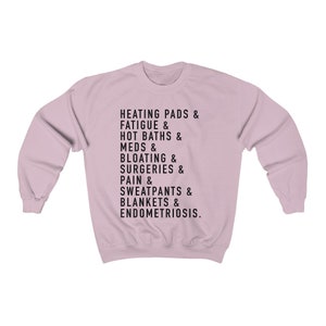 May include: A light pink sweatshirt with black text that reads "HEATING PADS & FATIGUE & HOT BATHS & MEDS & BLOATING & SURGERIES & PAIN & SWEATPANTS & BLANKETS & ENDOMETRIOSIS."