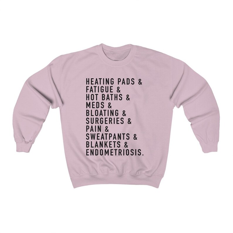 May include: A light pink sweatshirt with black text that reads "HEATING PADS & FATIGUE & HOT BATHS & MEDS & BLOATING & SURGERIES & PAIN & SWEATPANTS & BLANKETS & ENDOMETRIOSIS."