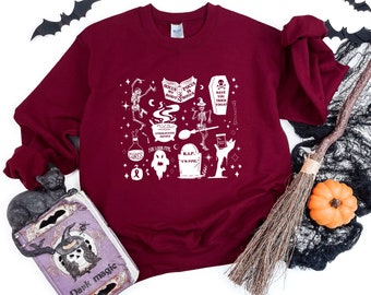 May include: A maroon sweatshirt with a white graphic print of a cauldron, a ghost, a tombstone, and other Halloween-themed imagery. The text on the graphic reads "Focus is my body, books is my soul", "Have you tried books?", "Undiluted Advice", "You look fine", "R.I.P. I'm fine", and "Curses".