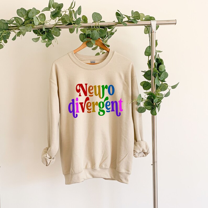 Może przedstawiać: Beżowa bluza z napisem "Neuro divergent" w kolorach tęczy.