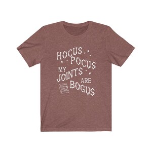 May include: A heather brown t-shirt with a white graphic that reads "Hocus Pocus My Joints Are Bogus" with a small book graphic that says "Chronic Pain Spell Book".