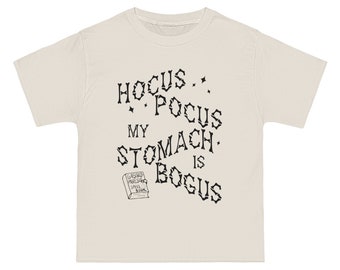 May include: A beige t-shirt with black text that reads "Hocus Pocus My Stomach Is Bogus". A small black and white illustration of a book with the text "Gas and Pressure Spell Book" is below the text.