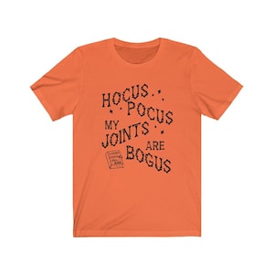 May include: Orange short-sleeved t-shirt with black text that reads "Hocus Pocus My Joints Are Bogus" and a small black and white illustration of a book with the text "Charms,  Finishes,  Spells,  Book".