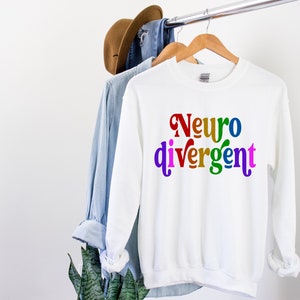 Może przedstawiać: Biała bluza z napisem "Neurodivergentny" w kolorach tęczy.