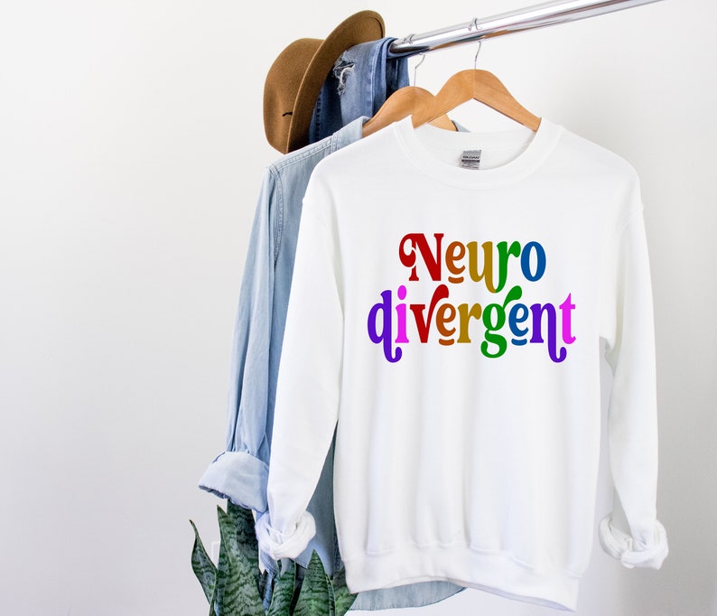 Może przedstawiać: Biała bluza z napisem "Neurodivergentny" w kolorach tęczy.