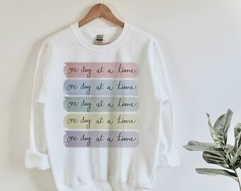 Puede incluir: Sudadera blanca con la frase "one day at a time" repetida cinco veces en un arcoíris de colores. Cada frase está escrita en un estilo manuscrito y se encuentra en una franja horizontal separada.