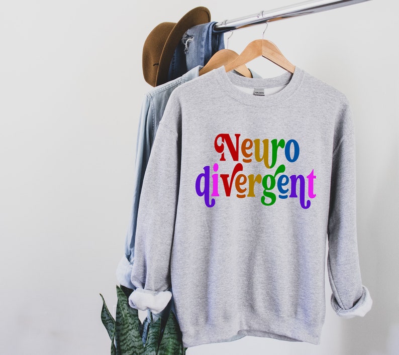 Może przedstawiać: Szara bluza z napisem "Neurodivergentny" w kolorach tęczy.