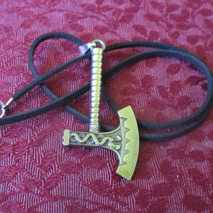 May include: A silver axe pendant with intricate details hangs on a black leather cord. The axe has a braided handle and a decorative design on the blade.
