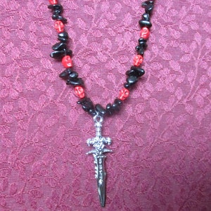 May include: A black and red beaded necklace with a silver sword pendant. The necklace is made of small, irregular beads and the sword pendant has a detailed design.