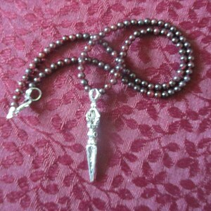 May include: A necklace featuring a strand of small, dark red beads. A silver-colored pendant with intricate details hangs from the center. The necklace is displayed on a textured, burgundy-colored surface.