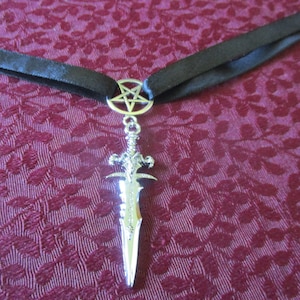 May include: A silver pendant necklace with a black ribbon. The pendant features a silver sword with a pentagram at the top.