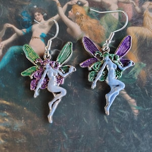 May include: A pair of fairy earrings with silver hooks. Each earring features a light blue fairy with purple and green wings. The background is a painting of nude women. The earrings are a whimsical accessory.