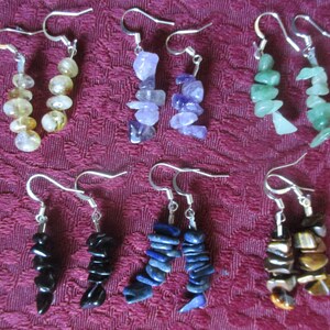 May include: A set of 8 pairs of gemstone earrings. Each pair features a different color of gemstone, including clear, yellow, purple, green, black, blue, and brown. The earrings are made with silver hooks.
