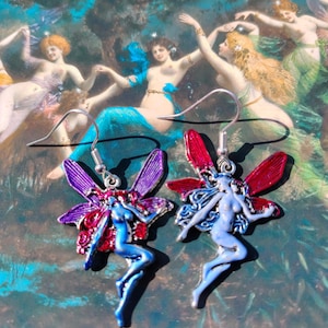 May include: A pair of fairy earrings. Each earring features a blue fairy with outstretched arms and red or purple wings. The background is a painting of dancing figures in a forest setting. The earrings are made of metal.