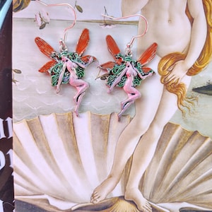 May include: A pair of fairy earrings with red and green wings and pink bodies. The earrings are hanging in front of a print of a painting depicting a woman in a seashell.