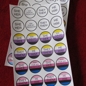 May include: A sheet of stickers with various pronoun options, including "she/her", "he/him", "they/them", and "ask for my pronouns". The stickers are in different colors, including yellow, purple, pink, blue, and black.
