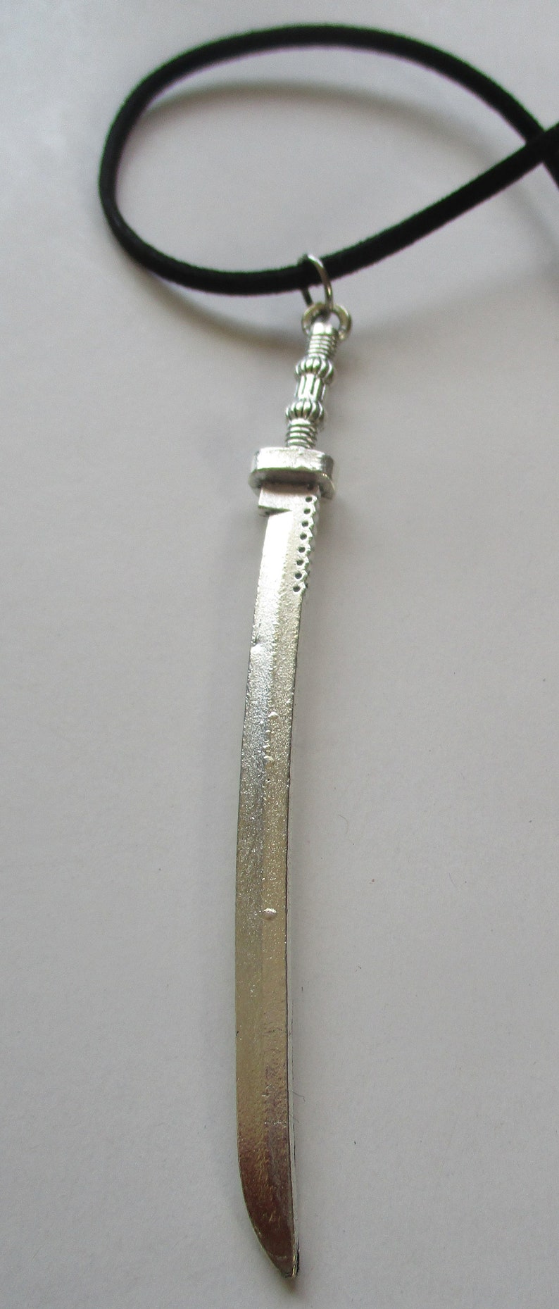 May include: A silver-colored sword pendant necklace with a black cord. The sword has a detailed hilt and a curved blade.
