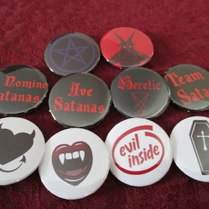 May include: Ten round black and white buttons with various satanic and gothic imagery. The buttons feature images of a pentagram, a devil, a coffin, a heart with horns, a set of fangs, and text such as "Ave Satanas", "In Nomine Satanas", "Team Satan", and "Heretic".