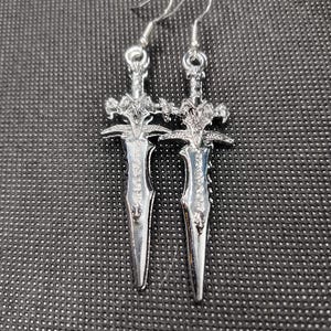 May include: A pair of silver sword earrings. The swords have intricate details and are hanging from silver hooks.