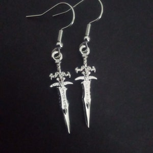 May include: A pair of silver-colored earrings featuring a sword design. Each earring has a hook for pierced ears and a detailed sword pendant. The swords have intricate designs on the hilt and blade, with a pointed tip.
