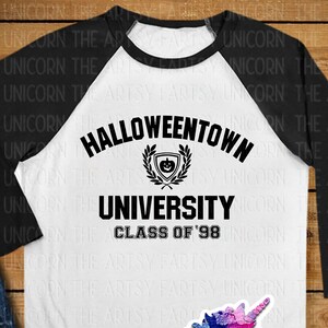 May include: A white and black baseball tee shirt with a black graphic that says "Halloweentown University Class of '98". The graphic includes a crest with a pumpkin inside. The shirt is laying on a wooden surface with a pair of black high-top sneakers and a pair of blue jeans.