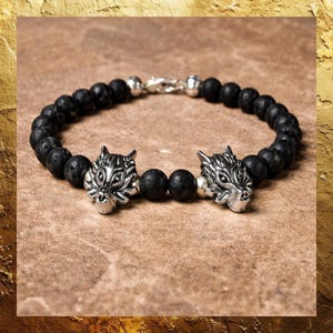 May include: A black beaded bracelet featuring two silver wolf head charms. The bracelet is made of small, round, black lava beads and has a silver clasp. The wolf heads are detailed with intricate facial features.