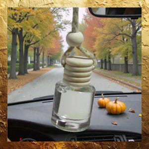 May include: A clear glass car air freshener with a wooden top and a hanging rope. The air freshener is hanging in front of a scenic autumn road with orange and yellow trees. Two small pumpkins sit on the dashboard.