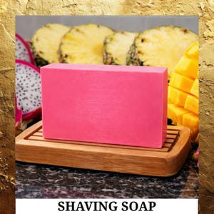 May include: A rectangular pink shaving soap bar rests on a bamboo soap dish. Slices of pineapple, dragon fruit, and mango are in the background. The text "SHAVING SOAP" is at the bottom of the image.