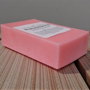 May include: A rectangular pink bar of scented soap with a white label. The soap is sitting on a wooden surface. The label reads "PERSEPHONE SCENTED SOAP".