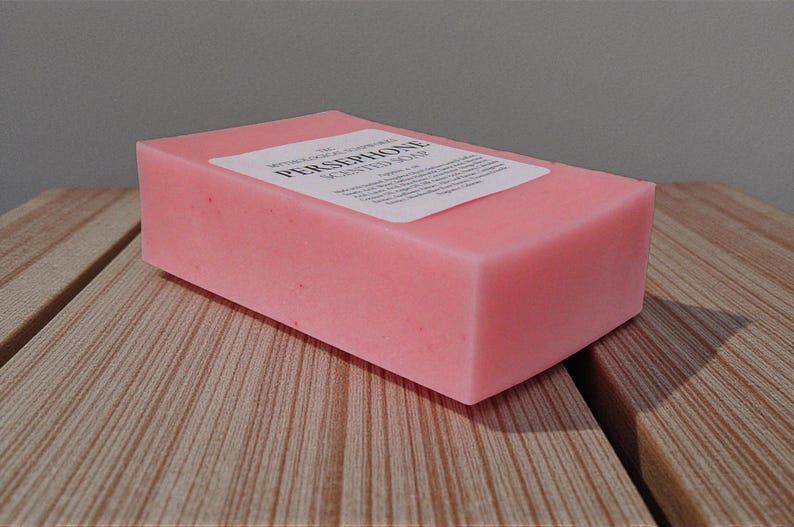 May include: A rectangular pink bar of scented soap with a white label. The soap is sitting on a wooden surface. The label reads "PERSEPHONE SCENTED SOAP".
