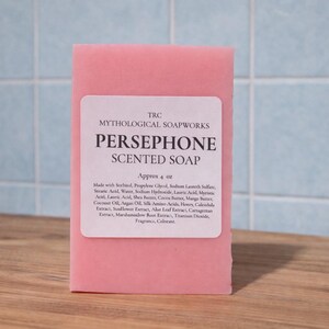 May include: A pink bar of Persephone scented soap from Mythological Soapworks. The soap has a white label with the product name and ingredients. The bar is approximately 4 oz. and sits on a wooden surface against a blue tiled background.